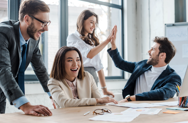 A group of excited colleagues celebrate a success in a bright office, with two team members giving a high-five.