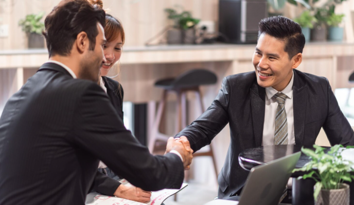 Three professionals in business attire shake hands and smile during a meeting in a modern office or cafe setting.