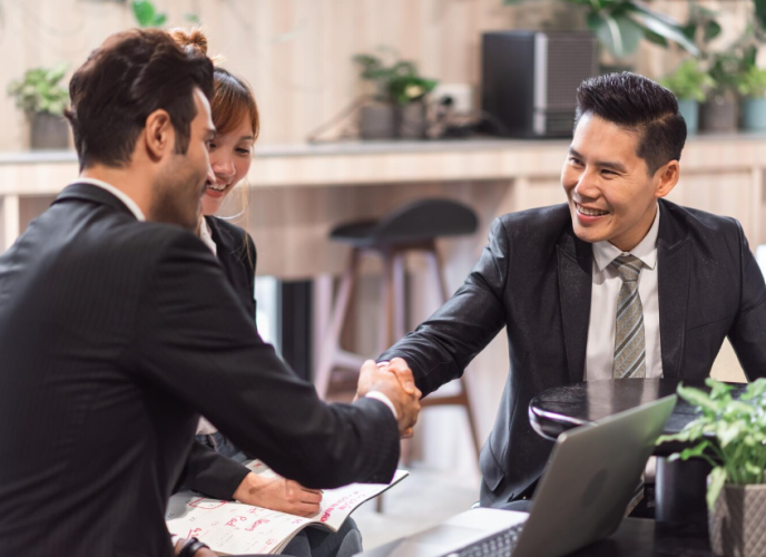 Three professionals in business attire shake hands and smile during a meeting in a modern office or cafe setting.